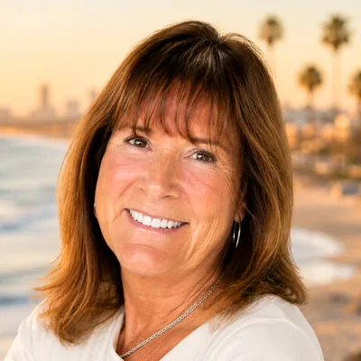Portrait of a smiling woman with shoulder-length auburn hair, a blurred beach and city skyline at sunset behind her